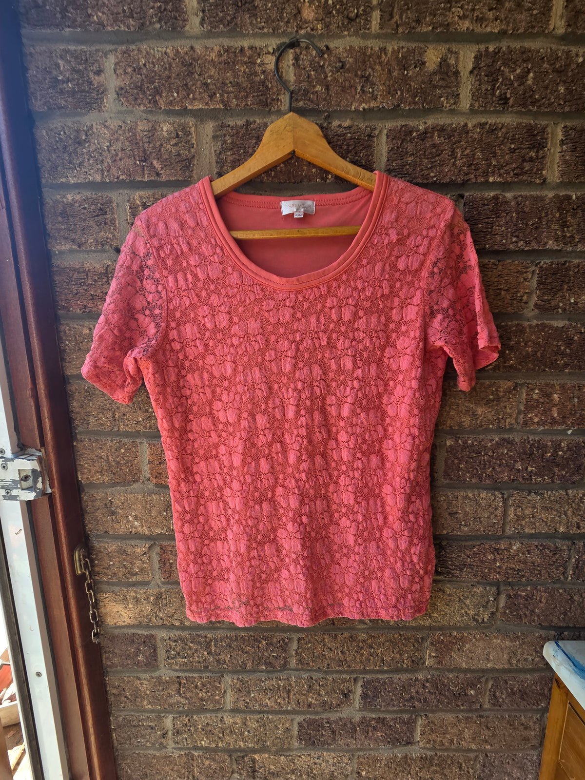 Paramour coral pink floral lace top on wooden hanger against brick wall, front view