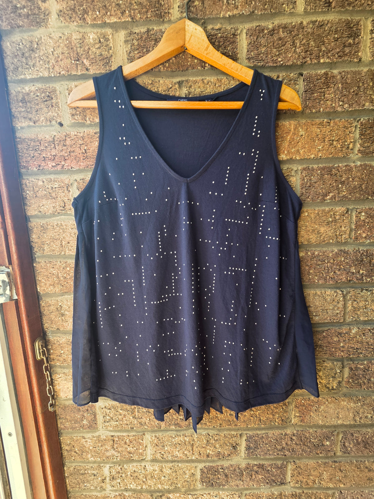 Next navy embellished vest top on wooden hanger against brick wall showing silver stud detail across front