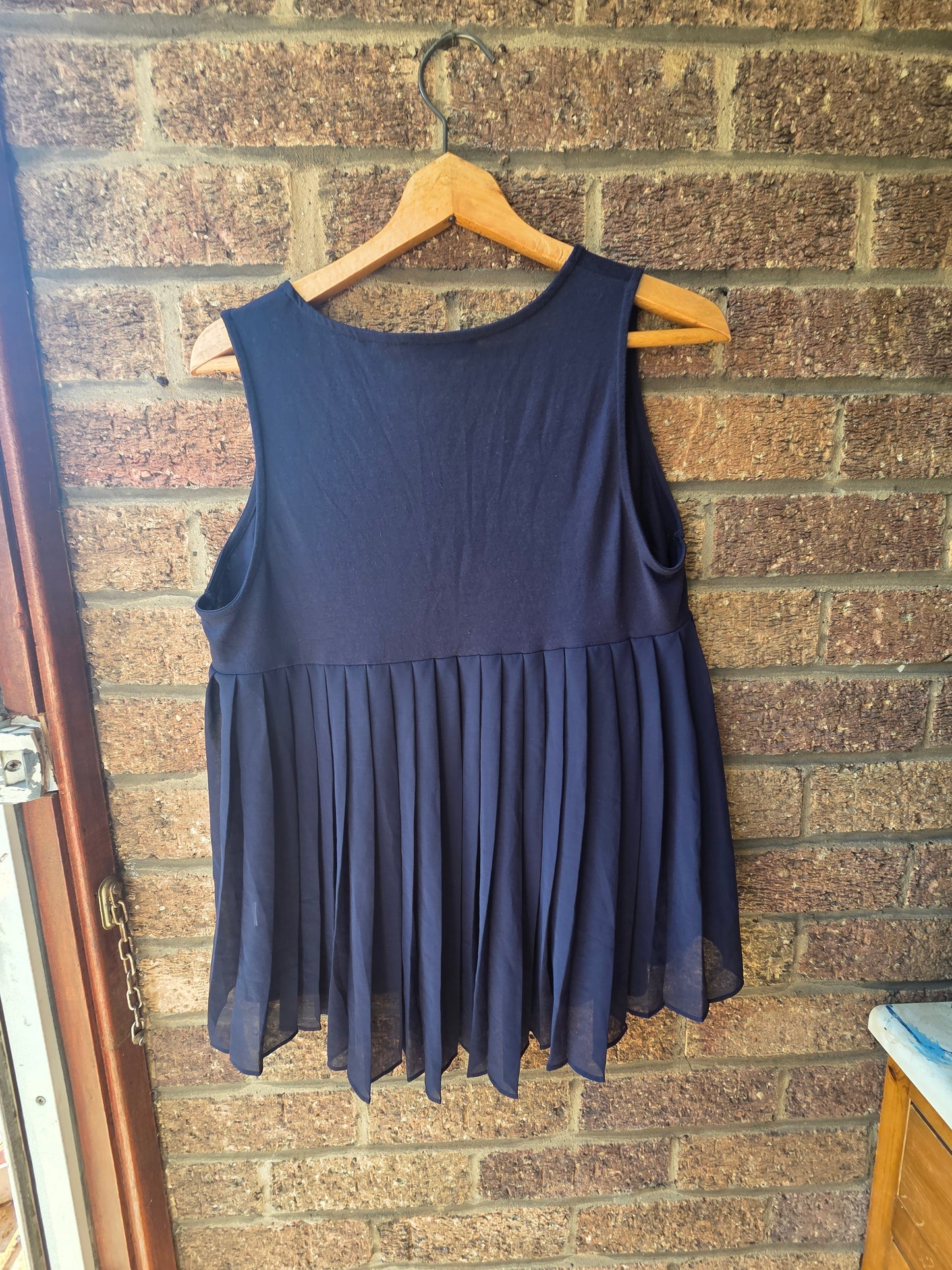 Next navy vest top on wooden hanger against brick wall showing pleated chiffon back panel with pointed hem